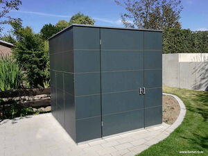 . The image shows a modern, sleek storage shed or garden structure with a flat roof and smooth, dark panels. It's located in a landscaped garden with paved pathways and surrounded by greenery and trees.