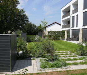 . The image shows a modern residential area featuring a white multi-story building with balconies and black shutters. In the foreground, there is a well-maintained garden with grass, bushes, and stepping stones. There are also pathways, patio furniture, and what appears to be outdoor storage sheds. Trees are present in the background.