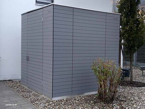 . Graues Gartenhaus mit moderner Verkleidung steht auf Kiesfläche neben Strauch und Baum.