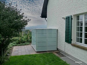 . Ein Gartenhaus neben einem Haus mit Fenster und grünen Fensterläden, umgeben von einem Garten und Bäumen.