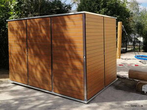 . Ein großer Holzschrank steht im Freien, umgeben von Bäumen und einem Spielplatz.