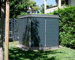 . The image shows a modern, rectangular outdoor shed with horizontal slats and a sleek design. It's situated on a grassy area, surrounded by trees and shrubs. The shed appears to be made of metal or a similar material and is likely used for storage in a garden or backyard setting.