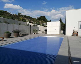 Ein moderner Garten mit Pool, Betonmauern, Pflanzen, Bänken und einem weißen Schuppen unter blauem Himmel.