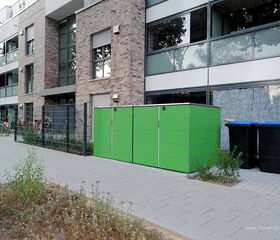 Moderne Gebäude mit Glas und Ziegel, grüner Schuppen, Mülltonnen, Fahrrad, Gehweg, Bäume.