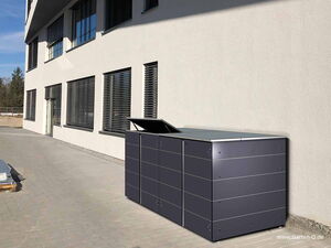 . The image depicts a modern outdoor storage unit or cabinet placed against the wall of a building. The unit has a sleek design, with a dark color and a horizontal panel pattern. One section of the storage unit appears to have its lid open. The building has multiple windows with horizontal blinds, and the scene is well-lit, suggesting daytime. There is also a paved area around the building.