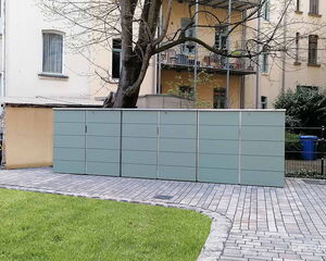 Ein grauer Mülltonnenunterstand vor einem Gebäude. Im Vordergrund eine gepflasterte Fläche und Rasen.