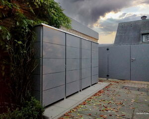 Grauer Gartenschrank neben einer Mauer, mit Pflanzen drumherum und einem bewölkten Himmel im Hintergrund.