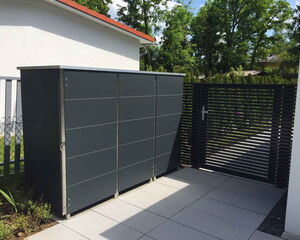 . The image shows a modern outdoor storage unit next to a metal gate. The flooring is comprised of concrete tiles, leading to a garden area with trees in the background. The environment appears well-maintained and is probably part of a residential or private outdoor space.