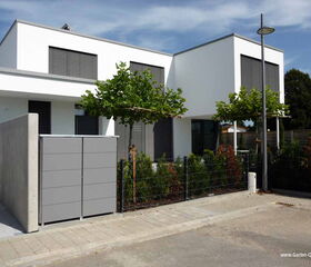 The image shows a modern residential building with a minimalist design. It has a flat roof and large windows with shutters. In the foreground, there's a paved pathway leading to the entrance and a gated area that may be used for storage or a utility enclosure. The property is landscaped with small trees, bushes, and a fence. There's a streetlamp on the sidewalk near the building.