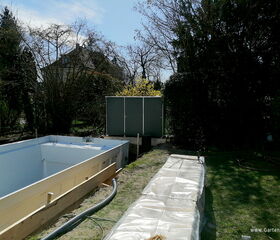Ein Garten mit leerem Pool, Gartenhäuschen und Bäumen. Bauarbeiten und Schläuche sichtbar.
