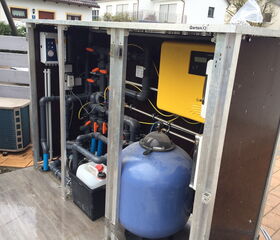 The image depicts an outdoor setup of pool equipment. It includes:

1. Various pipes and valves for water flow.
2. A filtration system, including a blue sand filter.
3. An electrical control unit (the yellow box).
4. A tank or container possibly for chemicals.
5. Wiring and connectors for electrical components. 

This arrangement is often used for maintaining pool circulation and filtration.