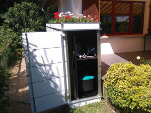 . The image shows an outdoor storage unit with its door open. Inside, you can see a hose reel and a container with a turquoise lid. The top of the storage unit has a planter with red and white flowers. The unit is situated next to a house with a window and a patio area, surrounded by greenery and bushes.
