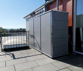 . Metallschrank auf Dachterrasse mit Geländer, daneben Lichtkuppel und Landschaft im Hintergrund.