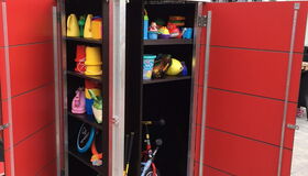 . The image shows a red cabinet with open doors containing various toys and equipment. Inside, there are plastic toy buckets, a tricycle, a colorful container, and other toys, likely meant for children's outdoor play. A mesh structure loaded with rocks is seen on top of the cabinet.