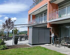 Haus mit Balkon, Terrasse, Garten und grauem Schuppen. Eine Person steht daneben.