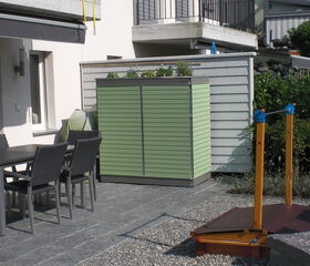 . The image shows a patio setting. Here's an array of what's visible:

1. Outdoor dining table with chairs
2. Green storage unit or cabinet
3. Wooden play structure or part of a climbing frame
4. Paved and pebbled ground
5. Building exterior with windows
6. Balcony with railing
7. Plants and shrubs nearby