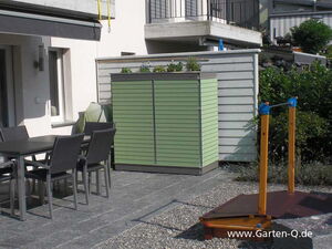 . The image shows a patio area with several elements:

1. A dining table with chairs made of a dark material.
2. A green garden storage unit with a planter on top, containing some plants.
3. The exterior of a house or building with windows and a railing above.
4. A small wooden structure with strings and blue elements, possibly part of a game or garden decor, on the right.
5. Paved and gravel ground surfaces.
6. Nearby greenery and shrubs.

Overall, it appears to be an outdoor living area with functional and decorative elements.