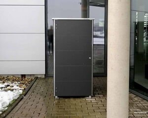 The image shows a black rectangular outdoor storage box or cabinet placed on a paved area near a building. It's surrounded by parts of a modern building structure, some rocks, and patches of snow.