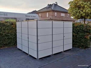. Ein moderner, grauer Gartenschrank auf einem gepflasterten Hof, umgeben von Büschen und einem Baum.