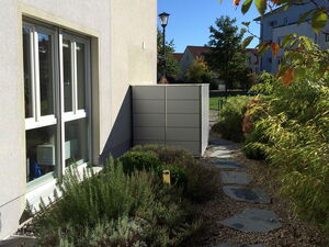 . In the image, there is a side view of a house with windows and a light-colored exterior wall. In front of the building, there's a small garden area with various green plants and shrubs. A stone path with irregularly shaped stones winds through the garden. There's a lamppost and a neatly organized fence or panel structure at the end of the path. Trees with green and yellow leaves are visible, indicating a sunny day. In the background, other residential buildings are visible.