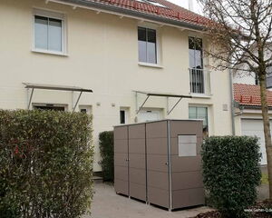 Ein modernes Haus mit zwei Eingängen, Fenstern und einem großen grauen Schuppen im Vorgarten, umgeben von Hecken.