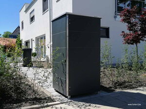 . Schwarzer Gartenschrank neben einem modernen weißen Gebäude, umgeben von Pflanzen und einem Bürgersteig.