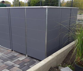 . The image shows a grey outdoor structure, possibly a storage unit or garden shed, on a paved area. There are plants nearby and a few people dressed in what appears to be work or gardening attire. The scene seems to involve some landscaping or garden work.