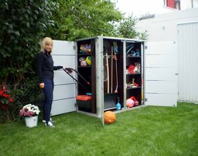 . The image shows a woman standing outside, next to an open outdoor storage shed. The shed contains various items, including gardening tools, a lawnmower, balls, and other assorted equipment. There are plants and a bucket of flowers nearby, and the scene is set in a grassy garden area.