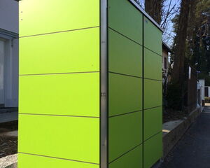 . The image depicts an electrical box or utility cabinet located outdoors. It features a modern design with bright green panels and metallic trim. It's situated on a paved area, likely near a residential or urban environment, with trees and a building visible in the background.