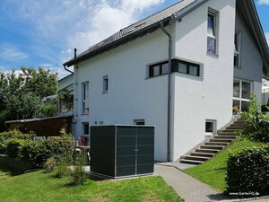. Ein modernes Haus mit Garten, Treppe, Rasen und einem großen schwarzen Schrank im Vordergrund.