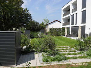 . Moderner Garten mit Rasen, Büschen, gepflasterten Wegen, zwei Schuppen, Terrasse mit Möbeln und Mehrfamilienhaus.