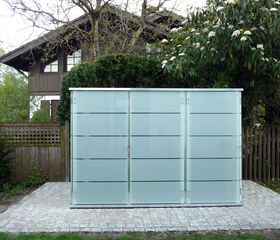. The image shows a modern, frosted glass structure, possibly a storage unit or shed, set on a stone or concrete patio. This is located in a garden with trees and foliage, and in the background, there is a wooden house partially visible. The structure contrasts with the natural setting around it.
