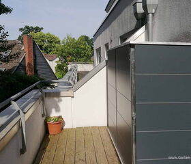 Eine Terrasse mit Holzbelag, Blumenkübel, Geländer und einem grauen Schrank neben einem Gebäude.