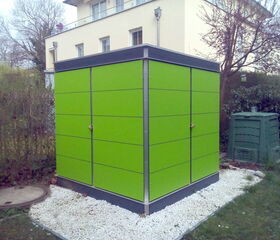 . The image shows a green structure in a garden setting. The structure appears to be a shed or a small building with a modern design, featuring smooth, bright green panels. It is surrounded by white gravel and the background includes a residential building, trees, and another green object that resembles a compost bin or storage unit.