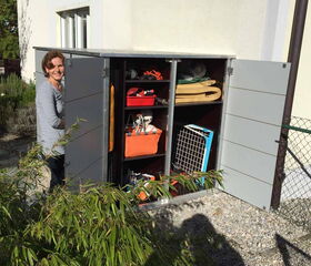 . The image shows an outdoor storage unit or shed with open doors, revealing shelves containing various items. These include storage boxes, bins, and possibly gardening tools or equipment. A person is standing next to the shed, looking inside. There are plants and a house wall in the background.