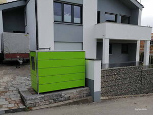 Grüne Mülltonnenbox vor einem modernen Gebäude mit Balkon und Anhänger auf gepflastertem Boden.