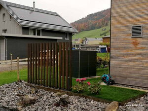 . Ein modernes Haus mit Solarpanels, daneben Garten mit Holzzaun, Pflanzen und Gartenschlauch.