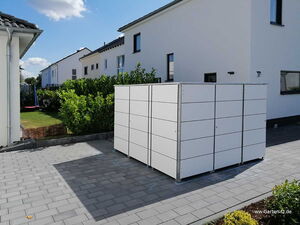. The image shows a paved outdoor area with a modern, modular storage unit in the center. The storage unit is composed of a grid-like structure with multiple rectangular panels. It is set against a background of residential houses with white walls and tiled roofs. There is greenery and a garden area nearby with bushes and grass. The sky is clear with a few clouds.