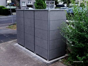 . Drei graue Mülltonnenboxen auf einem Gehweg neben grünen Büschen in einer städtischen Umgebung.