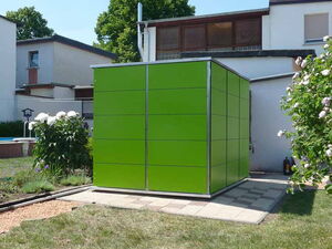 . The image shows a green cube-shaped structure made of panels situated in a garden area. It's placed on a paved surface and surrounded by grass, flowers, and bushes. There are houses in the background under a clear sky.