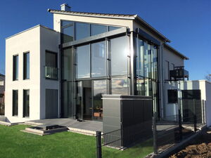 . The image shows a modern house with a combination of glass and solid walls. The design features large windows and an angular roof, with metal and glass elements providing a contemporary aesthetic. There's also a small grassy lawn in the foreground, surrounded by a fence. The overall setting appears bright and sunny.