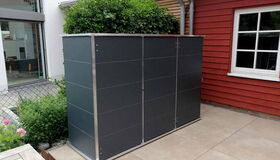 . Ein moderner Gartenschrank mit Pflanzendach steht auf einer Terrasse neben einem roten Haus.