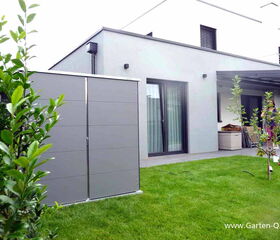 . The image shows a modern backyard with a sleek, gray garden storage unit on the left. There's a grassy lawn, a few plants, and a patio connected to a contemporary-looking house, featuring large glass doors and windows. The house has a minimalistic design with clean lines.