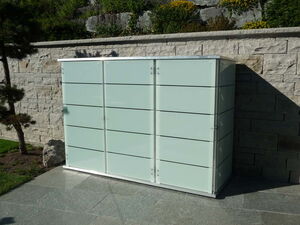 . The image shows a glass and metal storage cabinet outdoors, placed against a stone wall. The cabinet has a modern design with horizontal lines and is set on a tiled surface. There are plants and greenery around the area.