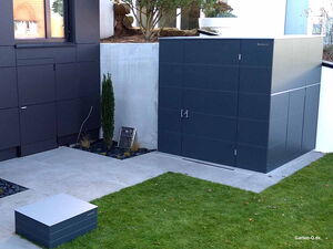 . Ein moderner, grauer Gartenschrank steht neben einem Gebäude auf einem befestigten Bereich mit angrenzender Rasenfläche.