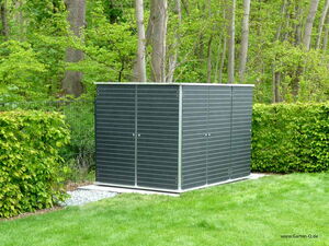 . The image shows a dark-colored garden storage shed located in a lush, green garden. The shed is surrounded by trees and hedges, and it sits on a grassy area with a small patch of gravel at its base. The shed has a simple, modern design with horizontal lines.