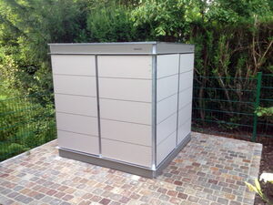 . The image shows a small, modern outdoor storage shed. It is rectangular with a flat roof and made of metal or similar material, featuring a minimalist design. The shed is situated on a paved area with a green fence and trees in the background.