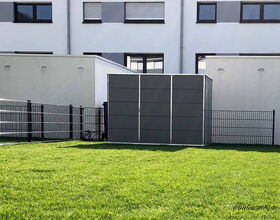 The image shows a modern-looking outdoor storage unit or shed, situated in a fenced area with a grassy lawn. Behind it, there is a residential building with multiple windows. The shed appears to be made of grey panels.
