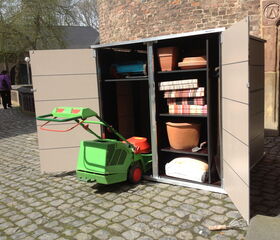 . The image shows an outdoor storage shed with its doors open. Inside the shed, there's a variety of gardening equipment and supplies, including:

1. A green lawn mower placed outside the shed.
2. Pots and planters on the shelves.
3. Some folded cushions or fabric items.
4. A bag of gardening soil or fertilizer.
5. A small hand shovel on one of the shelves.

The shed is located on a cobblestone surface and there are a few people in the background.