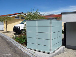 . The image features an outdoor setting with a modern storage unit or shed. It's located in a residential area, next to a driveway or parking area with a vehicle visible in the background. The storage unit has a sleek design with horizontal lines across its surface, and there are plants and landscaping around it. The environment appears to be neat and well-maintained.